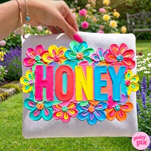 May include: A white pillow with the word "HONEY" in colorful, raised lettering, surrounded by vibrant, textured flowers in shades of pink, yellow, blue, and orange. The pillow is held against a background of green grass and colorful flowers.