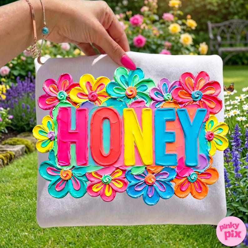 May include: A white pillow with the word "HONEY" in colorful, raised lettering, surrounded by vibrant, textured flowers in shades of pink, yellow, blue, and orange. The pillow is held against a background of green grass and colorful flowers.