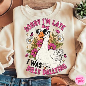K&ouml;nnte beinhalten: Cremefarbenes Sweatshirt mit einer Grafik einer Gans mit Sonnenbrille und rosa Schleife. Die Gans ist von rosa Blumen, G&auml;nsebl&uuml;mchen und Schmetterlingen umgeben. Der Text auf dem Sweatshirt lautet "SORRY I'M LATE I WAS DILLY DALLYING."