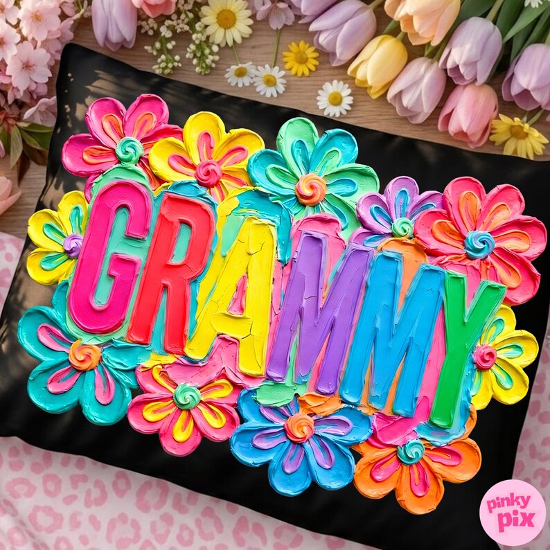 May include: Black pillow with the word "Grammy" in colorful, raised lettering, surrounded by vibrant, textured flowers in shades of pink, yellow, blue, and orange. The pillow is a decorative item.