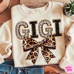May include: Cream-colored sweatshirt with the word "GIGI" in large, jeweled letters. The letters are outlined in gold and black, with clear rhinestones. A leopard print bow with gold studs is centered below the text.