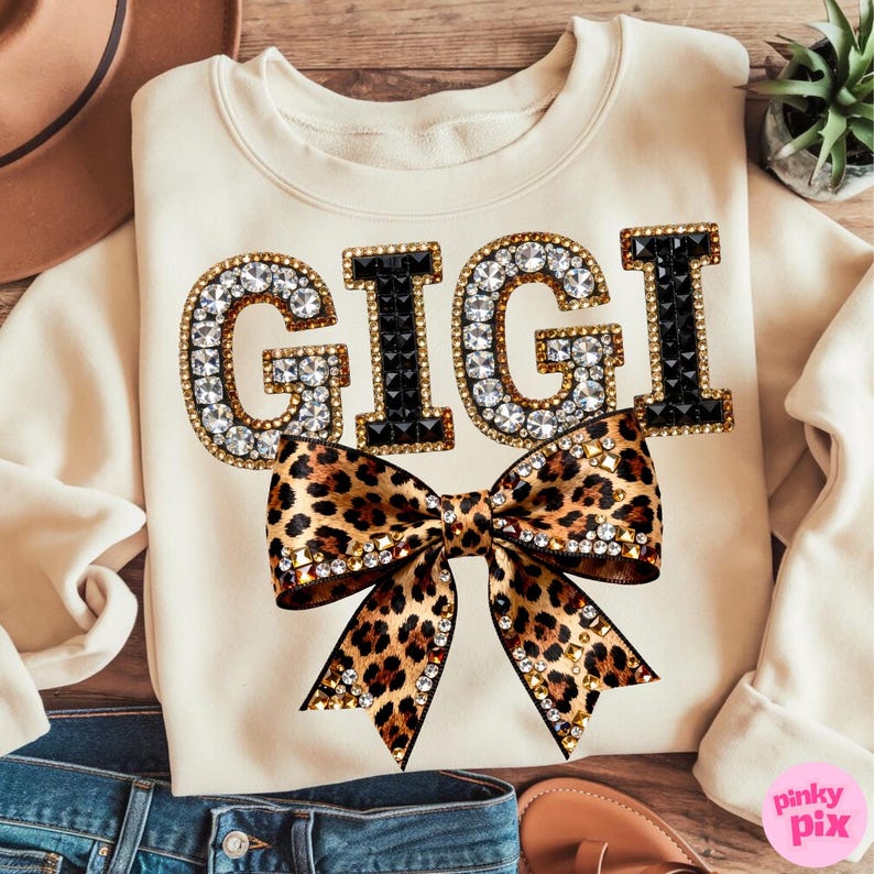 May include: Cream-colored sweatshirt with the word "GIGI" in large, jeweled letters. The letters are outlined in gold and black, with clear rhinestones. A leopard print bow with gold studs is centered below the text.
