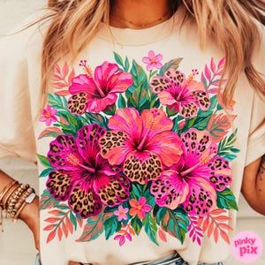 May include: A beige t-shirt featuring a vibrant floral design. The design includes large pink hibiscus flowers with leopard print centers, surrounded by green and orange leaves. The shirt has a crew neck and short sleeves.