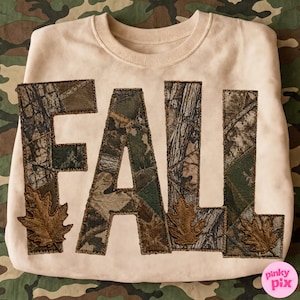 May include: A light beige crewneck sweatshirt featuring the word FALL in large, block-style letters. The letters are filled with a camouflage pattern and decorated with embroidered brown oak leaf appliques at the base of each letter.