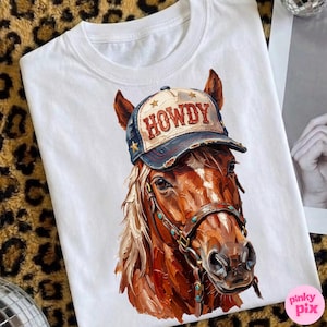 May include: White t-shirt featuring a detailed illustration of a brown horse wearing a blue and tan cap with the word "HOWDY" in red. The horse has a bridle with turquoise accents. The shirt is a casual wear item.