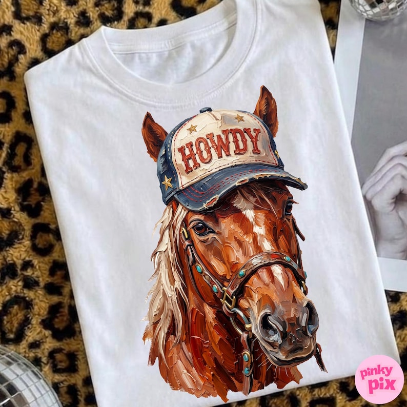 May include: White t-shirt featuring a detailed illustration of a brown horse wearing a blue and tan cap with the word "HOWDY" in red. The horse has a bridle with turquoise accents. The shirt is a casual wear item.