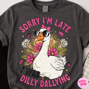 K&ouml;nnte beinhalten: Dunkelgraues Sweatshirt mit einer wei&szlig;en Gans, die eine Sonnenbrille und eine rosa Schleife tr&auml;gt. Die Gans ist von rosa und wei&szlig;en Blumen, Schmetterlingen und dem Text "SORRY I'M LATE I WAS DILLY DALLYING" in Rosa umgeben.