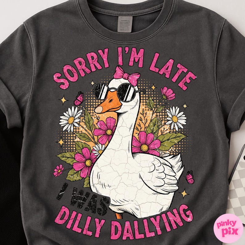 K&ouml;nnte beinhalten: Dunkelgraues Sweatshirt mit einer wei&szlig;en Gans, die eine Sonnenbrille und eine rosa Schleife tr&auml;gt. Die Gans ist von rosa und wei&szlig;en Blumen, Schmetterlingen und dem Text "SORRY I'M LATE I WAS DILLY DALLYING" in Rosa umgeben.