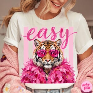 May include: White t-shirt featuring a tiger wearing pink sunglasses and a pink feather boa. The word "easy" is written in pink script above the tiger, with pink vertical stripes in the background.