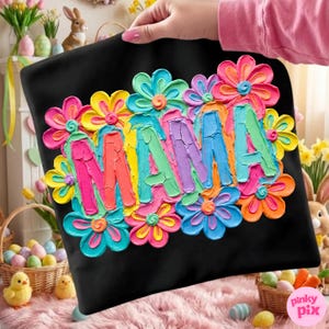 May include: Black tote bag with the word "MAMA" in colorful, textured letters surrounded by vibrant, layered flower designs in shades of pink, yellow, blue, and orange. The bag is held by a person with pink nail polish.