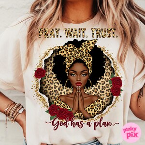 May include: A white t-shirt with a graphic design featuring a black woman with a leopard print headband and a gold glitter background. The text "Pray. Wait. Trust. God has a plan." is printed on the shirt.