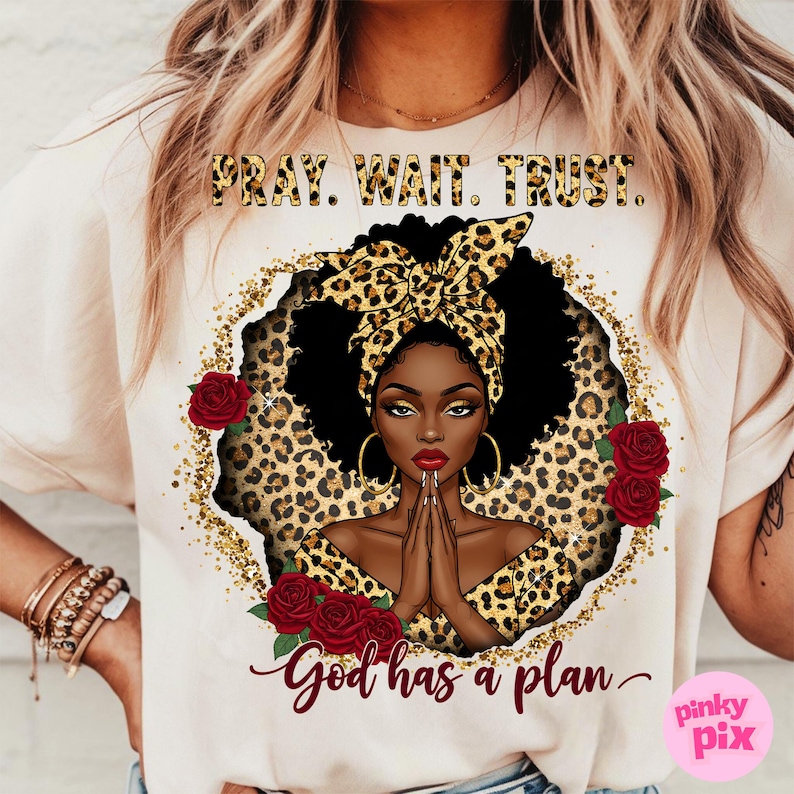 May include: A white t-shirt with a graphic design featuring a black woman with a leopard print headband and a gold glitter background. The text "Pray. Wait. Trust. God has a plan." is printed on the shirt.