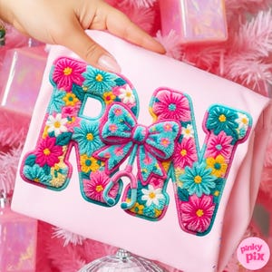 May include: A light pink pillow with the embroidered letters "RN" in a floral design. The letters are filled with pink, turquoise, yellow, and white flowers. A turquoise bow and stethoscope are between the letters.