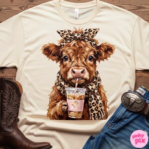 May include: Cream-colored t-shirt featuring a brown cow with a leopard print scarf and headband, sipping an iced coffee with a red and white striped straw. The cow is the central focus, with a detailed illustration style. The shirt is on a wooden surface.