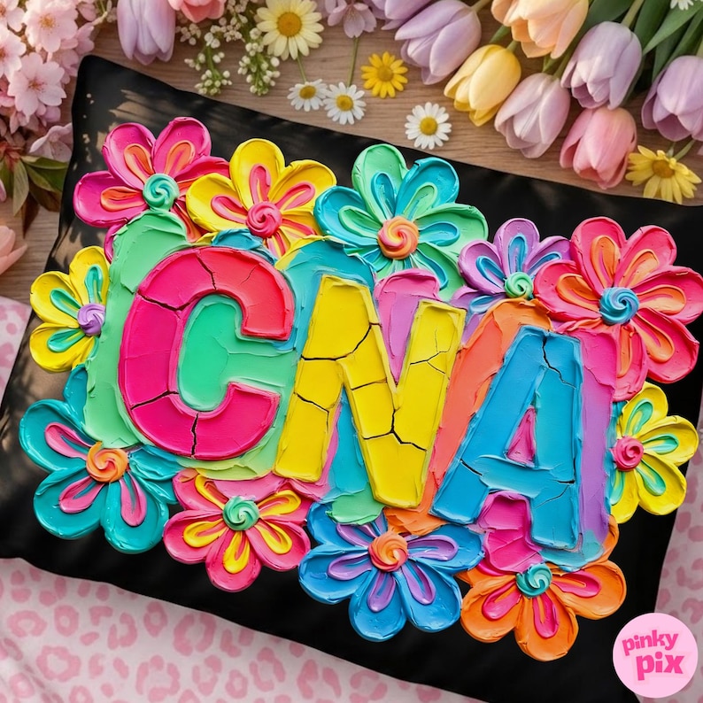 Op de afbeelding: Zwart kussen met kleurrijke, getextureerde bloemen en de letters "CNA" in heldere kleuren. De bloemen zijn in tinten roze, geel, blauw en oranje. Het kussen is omringd door bloemen.