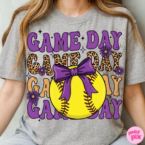 May include: Gray t-shirt with a softball graphic. The design features the words "GAME DAY" in purple, leopard print, and orange, with a yellow softball and purple bow. The shirt has a relaxed fit.