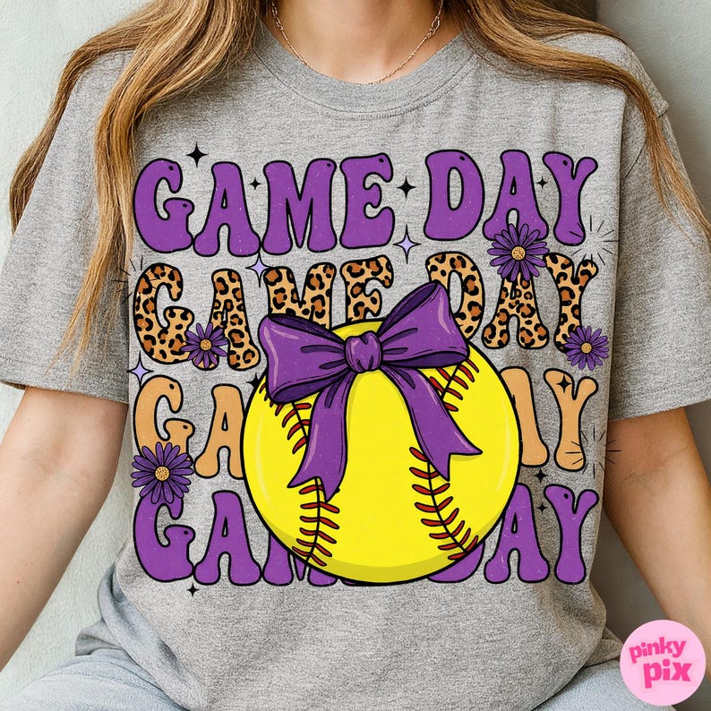 May include: Gray t-shirt with a softball graphic. The design features the words "GAME DAY" in purple, leopard print, and orange, with a yellow softball and purple bow. The shirt has a relaxed fit.
