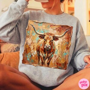 May include: A gray sweatshirt featuring a square, textured painting of a longhorn steer. The artwork uses a palette of browns, tans, and teal, with visible brushstrokes. The steer has large horns and a detailed face.