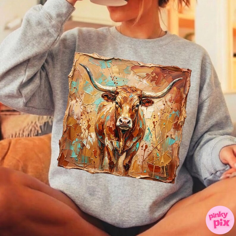 May include: A gray sweatshirt featuring a square, textured painting of a longhorn steer. The artwork uses a palette of browns, tans, and teal, with visible brushstrokes. The steer has large horns and a detailed face.