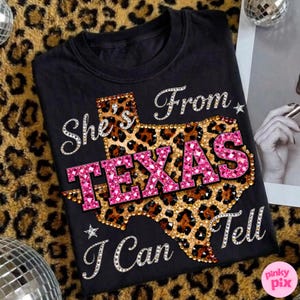 May include: Black t-shirt with the text "She's From Texas I Can Tell" in a rhinestone font. The word "TEXAS" is in pink rhinestones, and the state outline is leopard print.