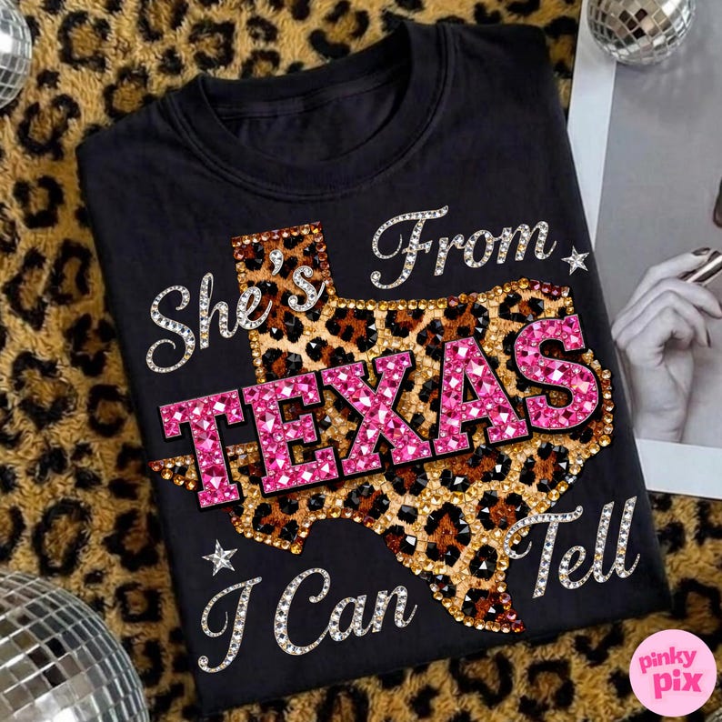 May include: Black t-shirt with the text "She's From Texas I Can Tell" in a rhinestone font. The word "TEXAS" is in pink rhinestones, and the state outline is leopard print.