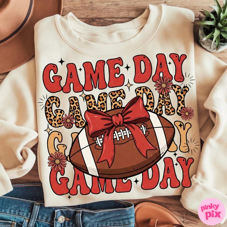 May include: Cream-colored sweatshirt with the text "GAME DAY" in red, leopard print, and yellow lettering. A football with a red bow is in the center. The design includes floral accents, stars, and a retro font.