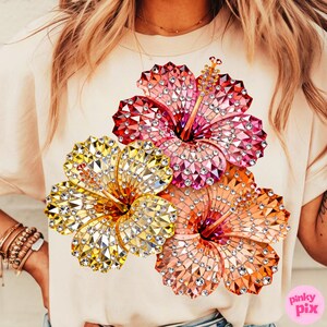 May include: A light beige t-shirt featuring a vibrant floral design. The design showcases three hibiscus flowers in shades of yellow, pink, and orange, embellished with sparkling crystals. The shirt has short sleeves and a round neckline.