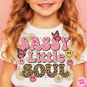 May include: White t-shirt with the words "SASSY Little SOUL" in pink, leopard print, and checkerboard lettering. The design includes butterflies, daisies, smiley faces, and stars. The shirt is for children.