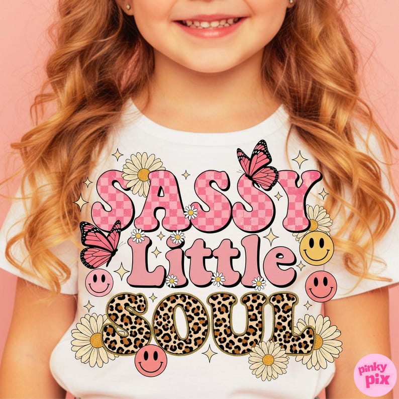 May include: White t-shirt with the words "SASSY Little SOUL" in pink, leopard print, and checkerboard lettering. The design includes butterflies, daisies, smiley faces, and stars. The shirt is for children.