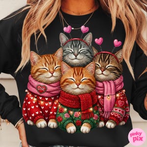 May include: Black sweatshirt featuring a graphic of four smiling cats. The cats wear sweaters, scarves, and heart-shaped headbands. The sweaters are red and green with heart and rose patterns. One scarf says "LOVE".