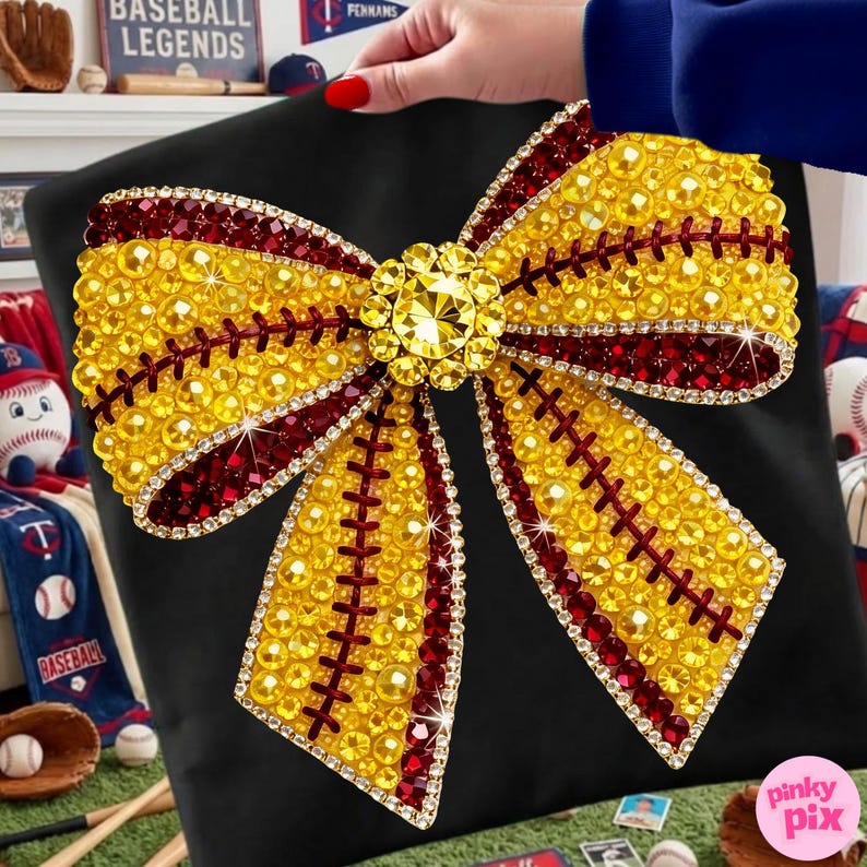 May include: A black cushion with a large, decorative bow made of yellow and burgundy gemstones. The bow has a central jewel and baseball-style stitching. This cushion is likely a decorative item for a sports-themed room or a fan's space.