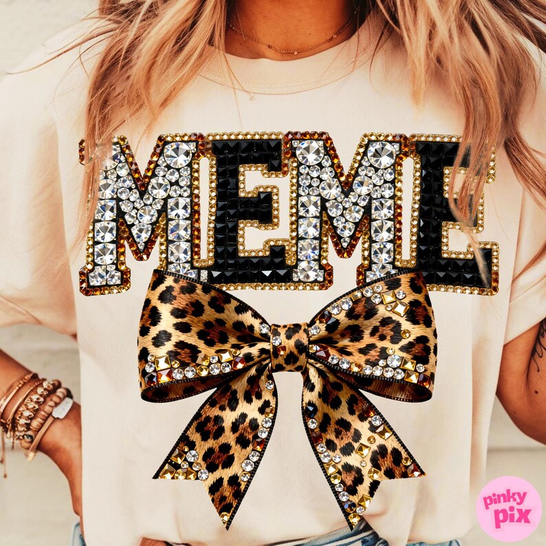 May include: A light beige t-shirt with the word "MEME" in large, jeweled letters. Below the text is a large leopard print bow with crystal embellishments. The shirt is a casual, stylish garment.