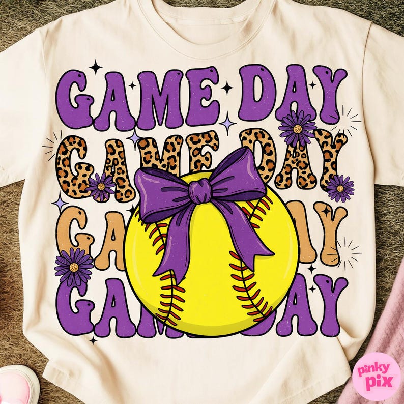 May include: Cream-colored t-shirt featuring the text "GAME DAY" in purple, leopard print, and tan. A yellow softball with a purple bow is in the center. Purple flowers and stars accent the design. The shirt is a casual wear item.
