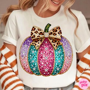 Op de afbeelding: Een wit T-shirt met korte mouwen en een grote afbeelding van een pompoen versierd met roze, paarse en turquoise strass-steentjes en witte sterren. De pompoen is bekroond met een strik met luipaardprint en goudkleurige randen.