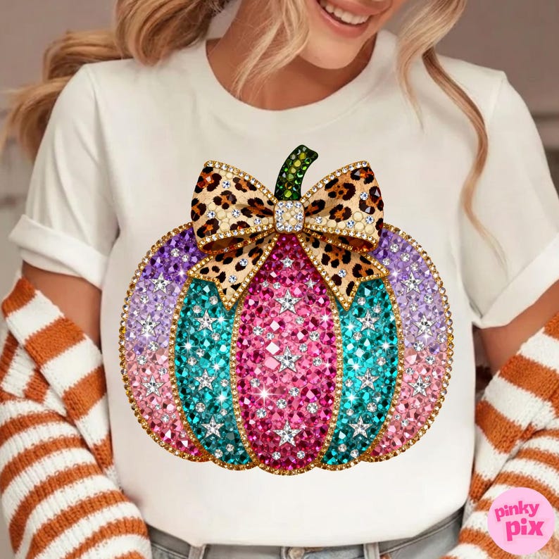 Op de afbeelding: Een wit T-shirt met korte mouwen en een grote afbeelding van een pompoen versierd met roze, paarse en turquoise strass-steentjes en witte sterren. De pompoen is bekroond met een strik met luipaardprint en goudkleurige randen.
