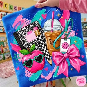 May include: A blue fabric square featuring a graphic print of a black and white composition notebook, a pencil with a checkered pattern, a pink apple wearing heart-shaped sunglasses, an iced coffee, a pink bow, and a lanyard with a smiley face tag.
