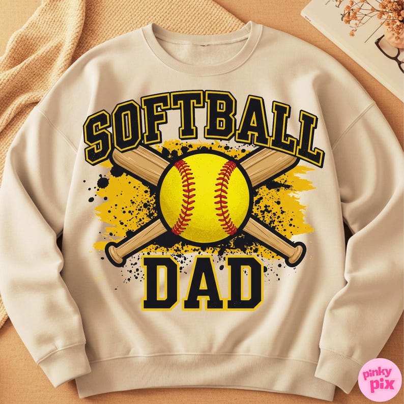 May include: Cream-colored sweatshirt with a softball graphic. The design features a yellow softball with red stitching, crossed wooden bats, and the words "SOFTBALL DAD" in black and yellow lettering.