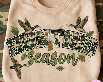 May include: A beige crewneck sweatshirt featuring the text Hunting Season in a camouflage pattern and dark green script. The design includes three flying mallard ducks and several oak leaves scattered around the bold, block-lettered text.