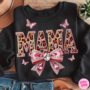 May include: Black sweatshirt with the word "MAMA" in leopard print, embellished with rhinestones. A pink bow with rhinestones and pink butterflies are also featured on the sweatshirt.