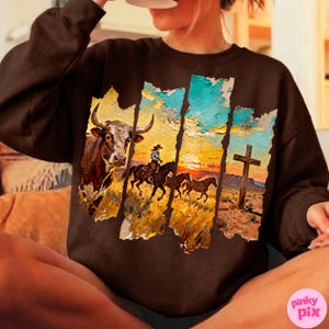 May include: A brown sweatshirt featuring a Western-themed graphic. The design includes a bull, a cowboy on horseback, and a cross against a sunset backdrop. The artwork has a textured, painted appearance.