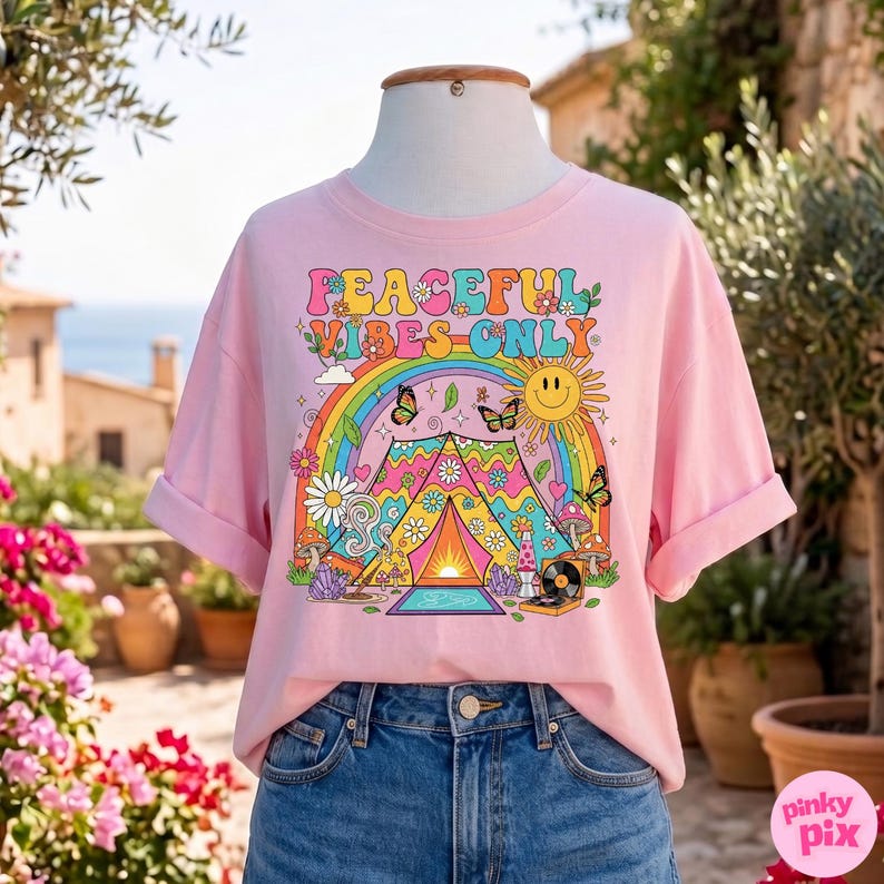 May include: A light pink t-shirt with rolled-up sleeves. The shirt features a colorful graphic with the text "PEACEFUL VIBES ONLY", a rainbow, butterflies, a sun, and a tent design. The shirt is displayed on a mannequin.