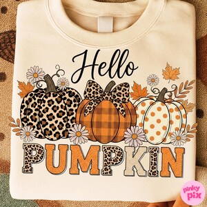 Op de afbeelding: Cr&egrave;mekleurige sweatshirt met de tekst "Hello Pumpkin" en drie ge&iuml;llustreerde pompoenen. De pompoenen hebben verschillende patronen, waaronder luipaardprint, ruitjes en stippen. Bloemen- en bladaccenten omringen de pompoenen.