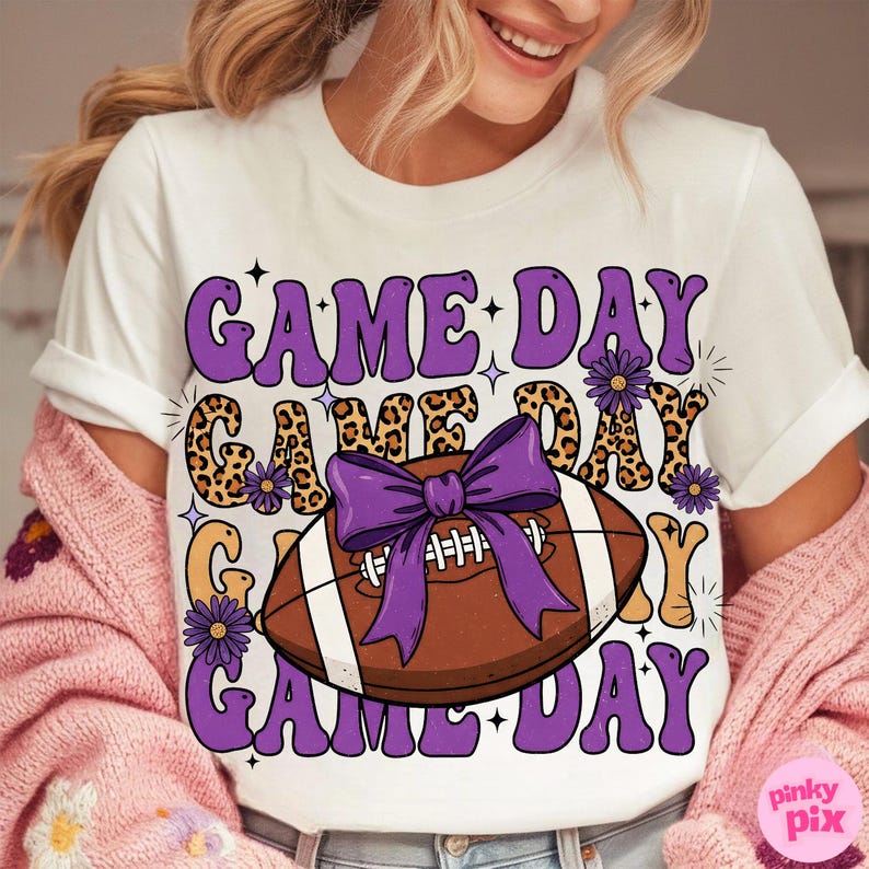May include: White t-shirt with a purple and leopard print "Game Day" design. The design includes a football with a purple bow and purple flowers. The shirt is a casual style.