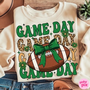 May include: Cream-colored sweatshirt with the text "GAME DAY" in green, leopard print, and yellow lettering. A brown football with a green bow is in the center. Green daisy accents are scattered around the text and football.