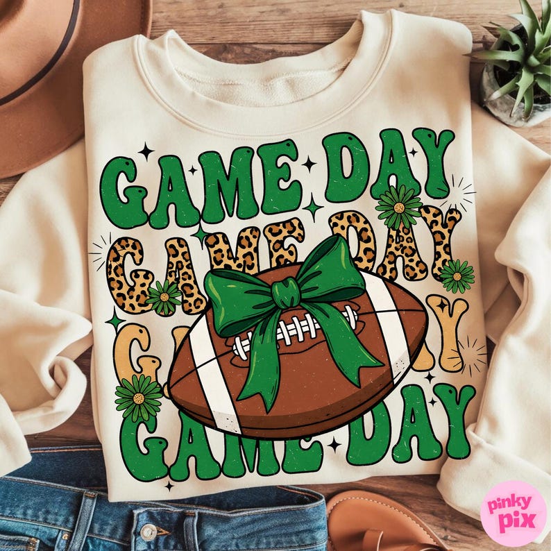 May include: Cream-colored sweatshirt with the text "GAME DAY" in green, leopard print, and yellow lettering. A brown football with a green bow is in the center. Green daisy accents are scattered around the text and football.