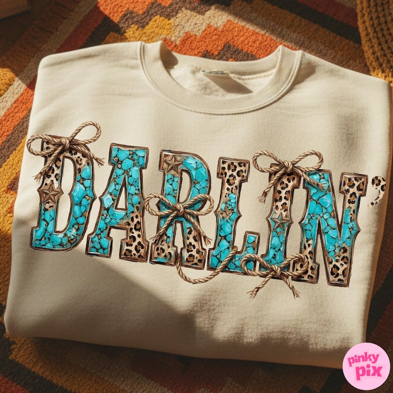 Pu&ograve; includere: Felpa beige con la scritta "DARLIN'" in un design turchese e leopardato. Le lettere sono decorate con fiocchi di corda e dettagli a stella, creando un'estetica di ispirazione western.