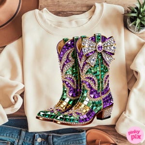 May include: Cream-colored sweatshirt with a graphic of jeweled cowboy boots. The boots are decorated with green, purple, and gold mosaic-style gems, and a large jeweled bow. The boots have a rhinestone-studded heel.
