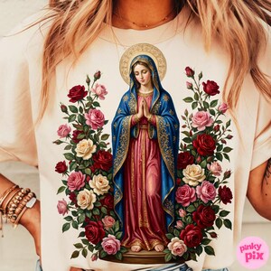 May include: Cream-colored t-shirt featuring a religious illustration of a figure in a blue cloak and red dress, surrounded by roses in shades of red, pink, and cream. The figure has a golden halo. The shirt has a Pinky Pix logo.