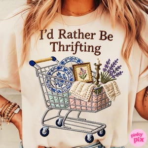 Puede incluir: Camiseta color crema con un diseño de carrito de compras bordado. El carrito está lleno de platos azules y blancos, un cuadro enmarcado, un candelabro, lavanda y una manta. El texto "I'd Rather Be Thrifting" está arriba.