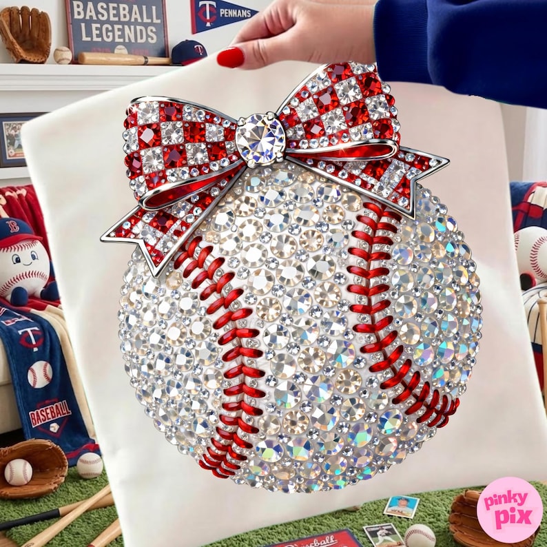 May include: A baseball-themed decorative pillow featuring a sparkling design. The baseball is covered in clear crystals with red stitching. A large bow with red and white checkered accents and a central crystal adorns the top. The pillow is on a white background.